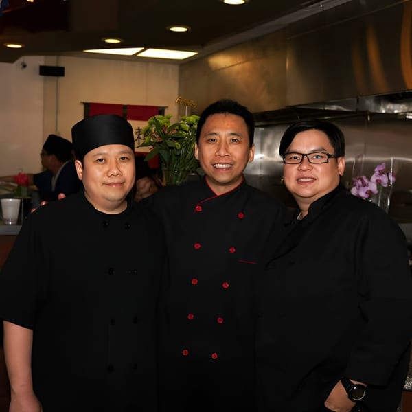 Three smiling Shinrai Japanese Bistro chefs in black uniforms pose together in the restaurant kitchen.