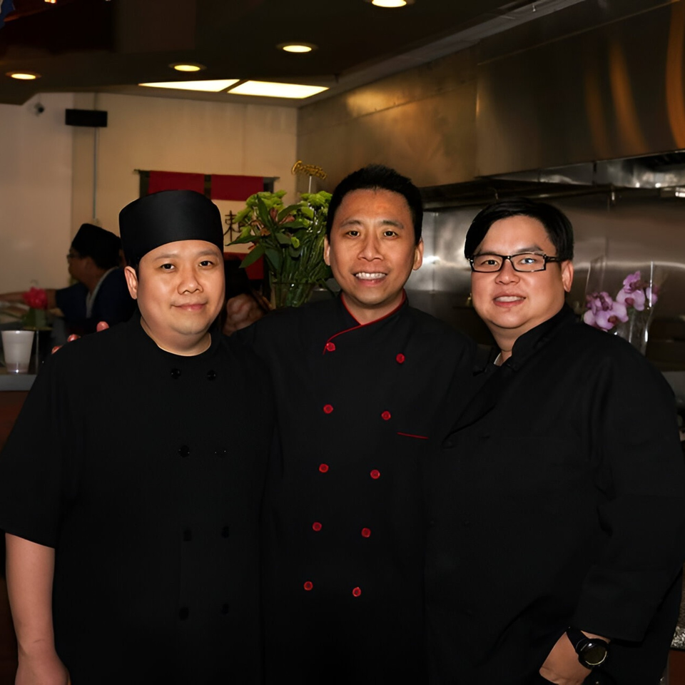Three smiling Shinrai Japanese Bistro chefs in black uniforms pose together in the restaurant kitchen.