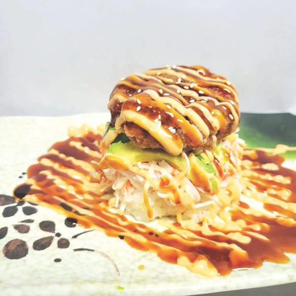 A Japanese sushi burger with crab, avocado, and a crispy patty, drizzled with sauces and sesame seeds.